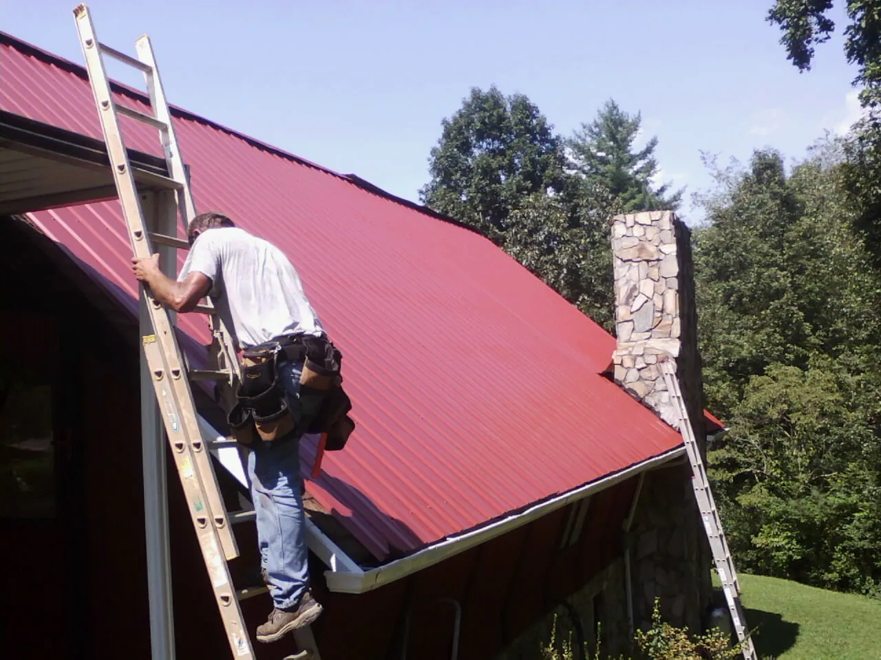 Roofer on a ladder beside a red metal roof and stone chimney — Soffit & Fascia Repair by Roofing Brothers in Newburyport